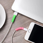 Lifestyle photo of green and pink spiral protectors on white charging cables connected to a laptop and smartphone on a wooden desk.